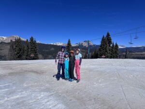 Five people in colorful ski outfits posing together on a snowy slope, with mountains and a chairlift in the background, under a bright blue sky.