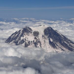 Snowy mountain peak with dark rocky faces, capped by snow, rising above a cloudy sea under a blue sky.