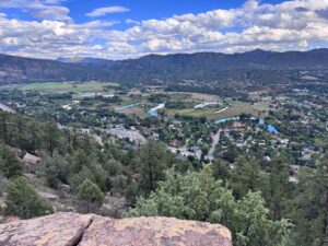 Panoramic valley view featuring a winding river, green fields, scattered houses, and forested hills beneath a blue sky with white clouds.